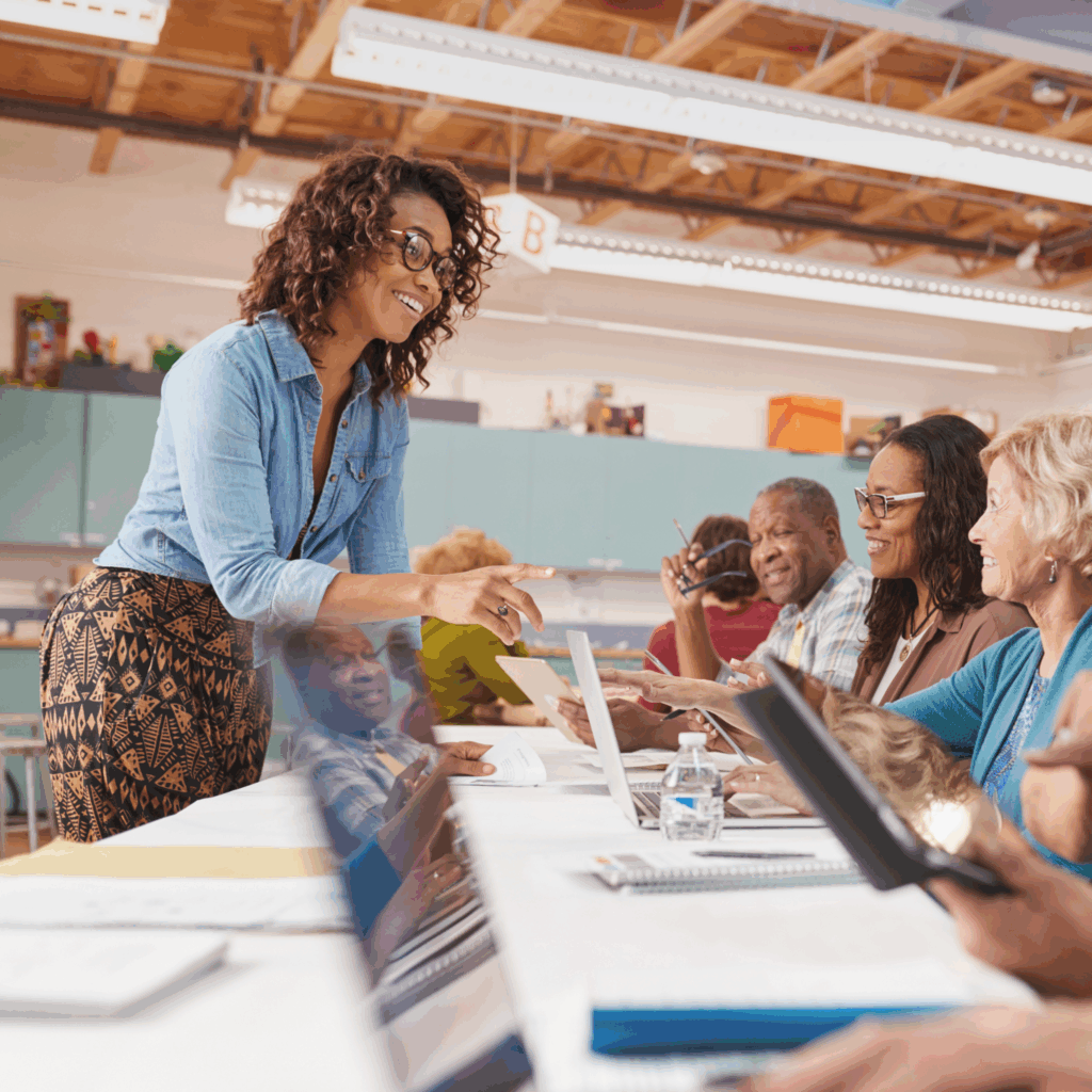Woman at a community event is standing at long table talking to others who are sitting in front of laptops